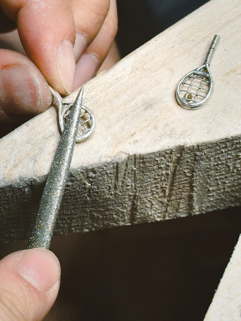 Close-up of hands making silver tennis racket charms on a wooden work bench